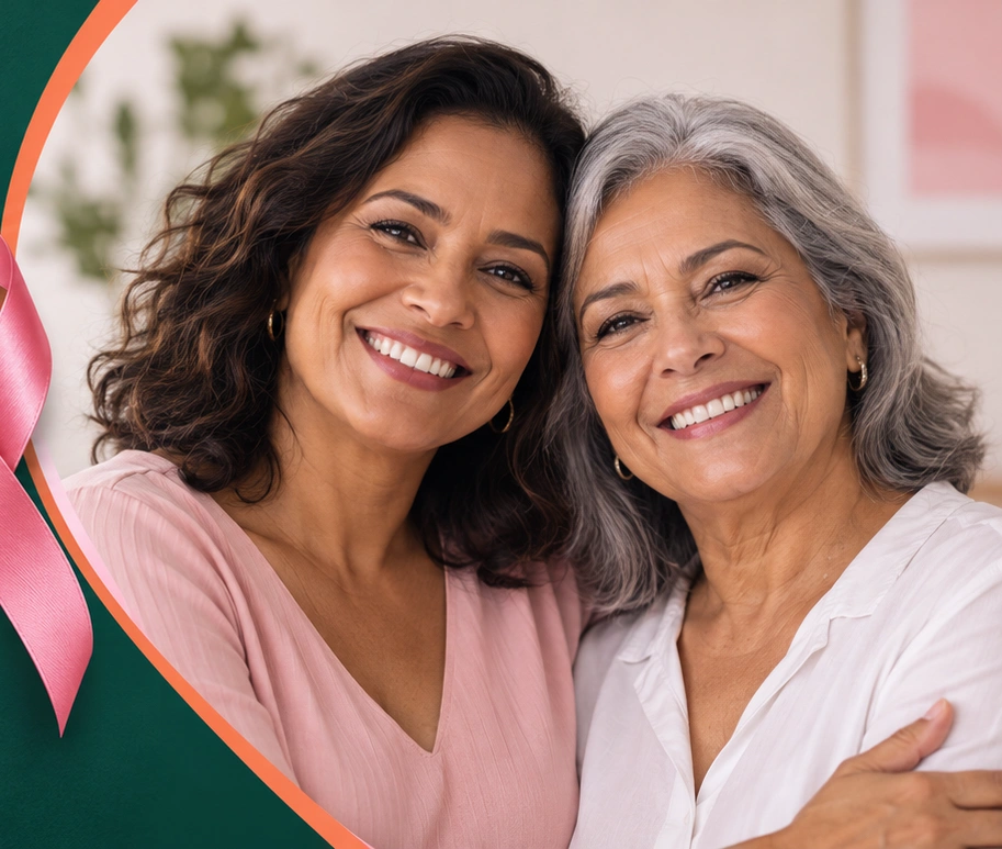 Duas mulheres sorrindo, abraçadas, ao lado do laço rosa da campanha Outubro Rosa.